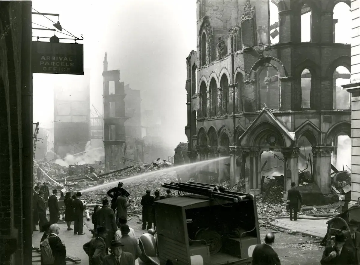 Firemen hosing water on a burning building following bomb damage with people onlooking