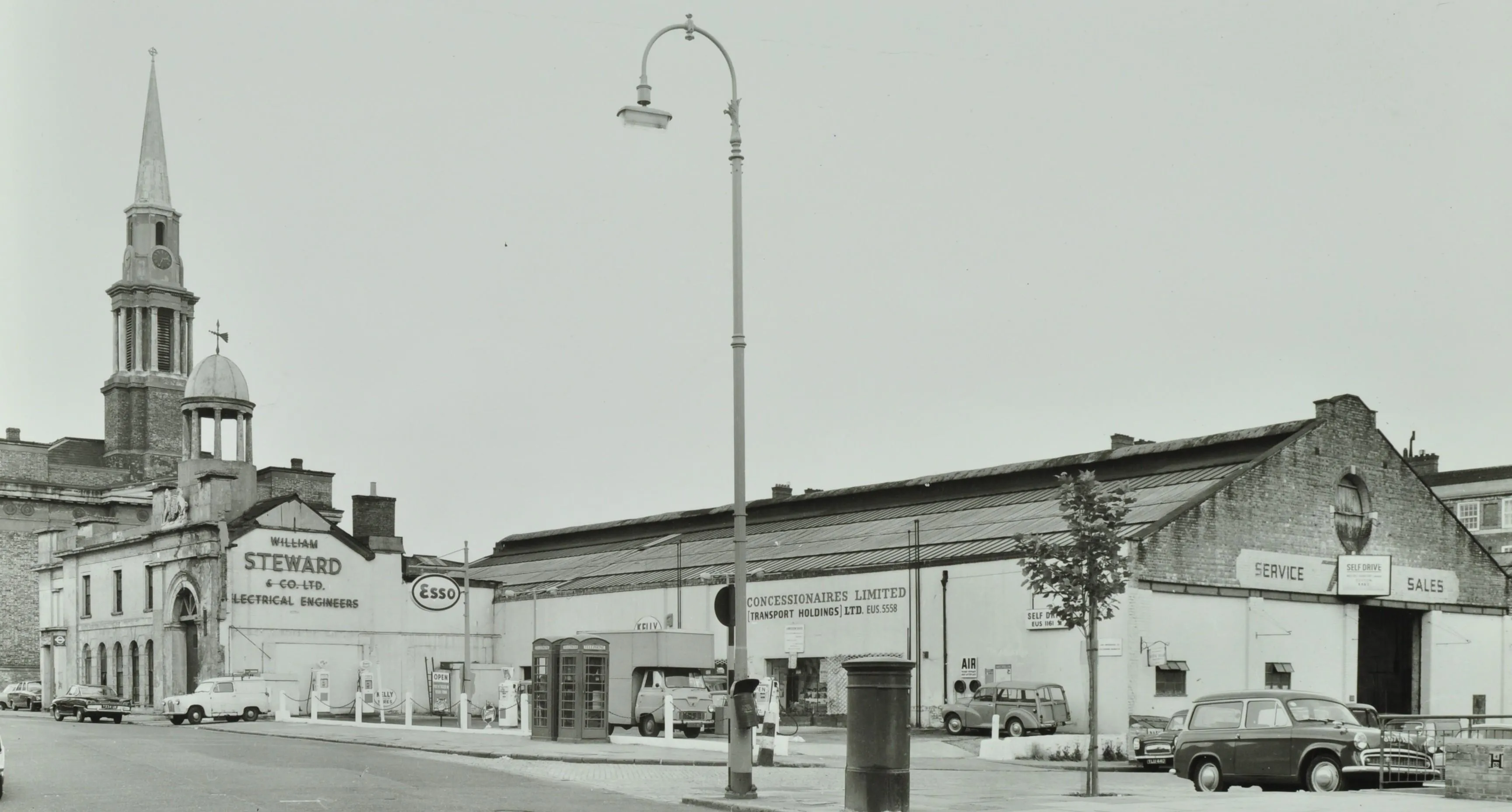 a low level garage building with church spire next door and cars, telephone and boxes and post box in the foreground